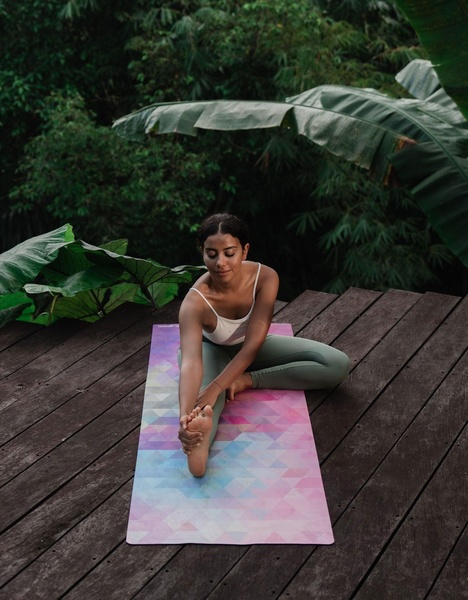 A high-angle shot of a woman performing a yoga stretch on a vibrant, geometric-patterned yoga mat set on a rustic wooden deck surrounded by lush jungle foliage.